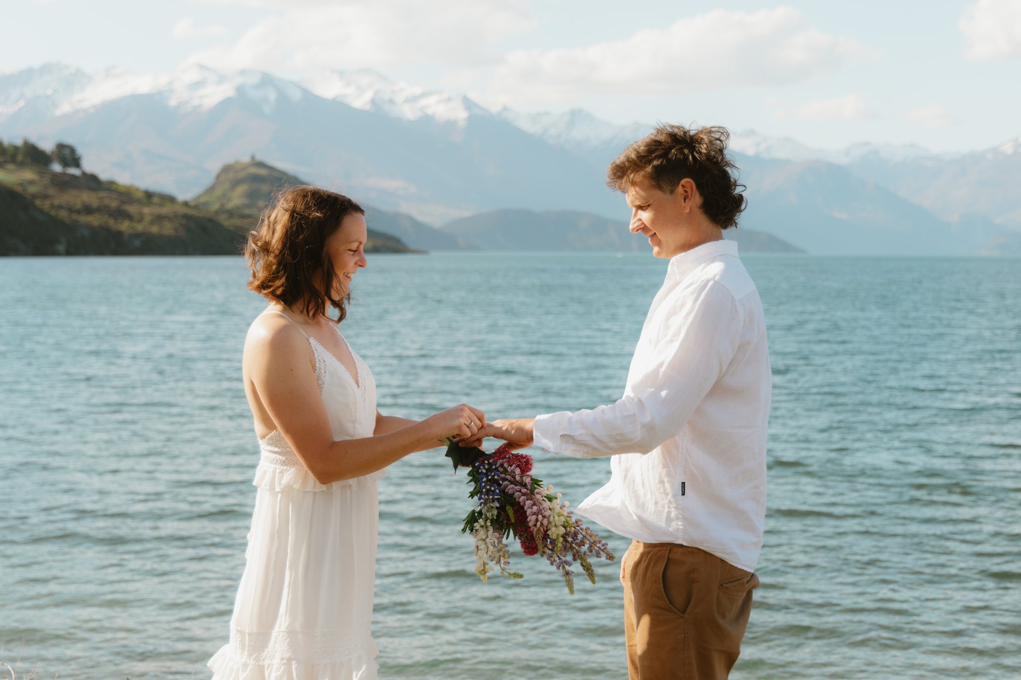 A couple in wedding outfits standing next to a lake with snowy mountains in the background and they are exchanging their weddings bands and the bride is holding a lupin bouquet.