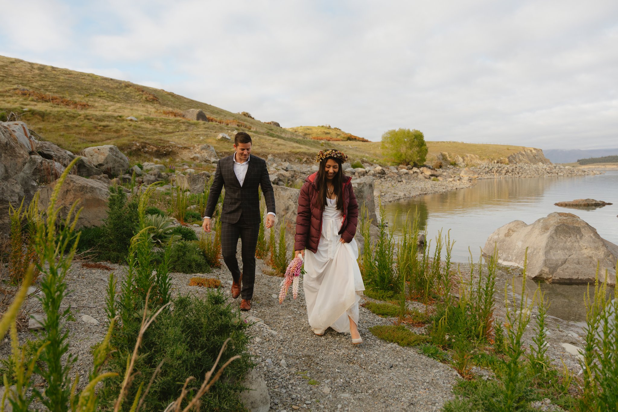 A couple walking alongside the water edge in between plants that are growing out of the rocky terrain. They are both in elopement outfits, the lady is wearing a wedding dress.